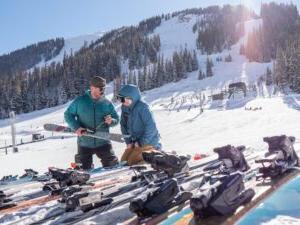 Summit High School teacher Thomas Lutke, right, adjusts a ski binding at Arapahoe Basin as student Makena Brendel looks on. Both are dressed in winter gear on a snowy slope, with ski equipment in the foreground.